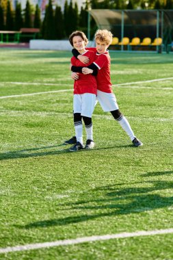 Futbol kıyafeti giyen iki genç çocuk yeşil futbol sahasında birbirlerine sevgiyle sarılıyorlar. Yüzleri birlikte kutlama yaparken mutluluk ve sportmenlik saçıyor..