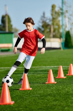 Genç bir çocuk futbol topunu enerjik bir şekilde turuncu konilerin etrafında tekmeliyor, çevikliğini ve yeteneğini sahada gösteriyor..