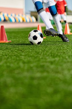 Spor kıyafeti giyen bir insan güneşli bir günde açık mavi gökyüzünün altında yeşil bir sahada futbol topunu tekmeliyor..