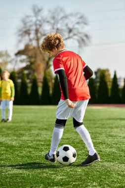 Kararlı bir ifadeyle genç bir çocuk parlak güneşin altındaki yemyeşil bir sahada futbol topunu tekmeliyor ve oyuna olan tutkusunu gösteriyor..