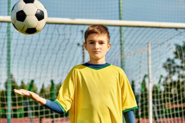 Genç bir çocuk yeşil bir futbol sahasında duruyor, elinde bir futbol topu tutarken heyecan saçıyor. Gözleri coşkuyla parlıyor, oynamaya hevesli ve yeteneklerini sergiliyor..