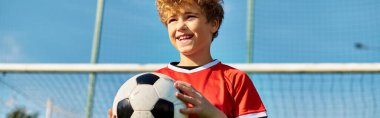 Geniş yeşil bir alanda duran genç bir çocuk, bir futbol topunu kararlılıkla kavrıyor. Önüne bakıyor, topa vurmaya ve heyecanlı bir oyun oynamaya hazır..