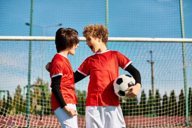 İki genç adam, birbirlerine yakın duruyorlar, ellerinde bir futbol topu var. Odaklanmış ve oyun için hazır görünüyorlar. Takım çalışması ve dostluk gösteriyorlar..