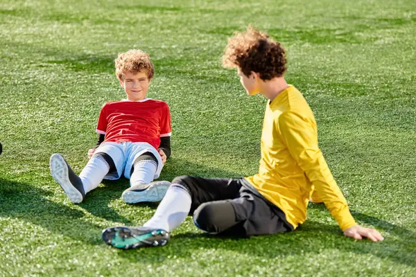 İki genç çocuk yemyeşil çim tarlasında enerjik bir şekilde futbol oynuyor. Top sürmekle, pas atmakla, topa vurmakla, yeteneklerini ve takım çalışmalarını sergilemekle meşguller..