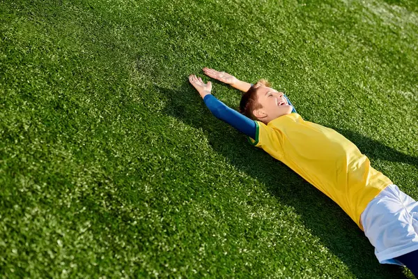 A young boy in a soccer uniform lies peacefully on the grass, staring at the sky with a smile on his face, lost in thoughts of the beautiful game.