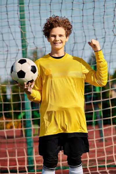 stock image A young boy stands in front of a soccer net, holding a soccer ball in his hands, with a look of determination on his face.