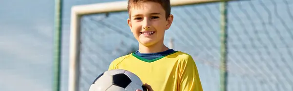 若い男の子が誇らしげに立って ゴールの前にサッカーボールを持っています 彼の目に決意を持って 彼はフィールド上のスタープレーヤーになることを夢見ています — ストック写真