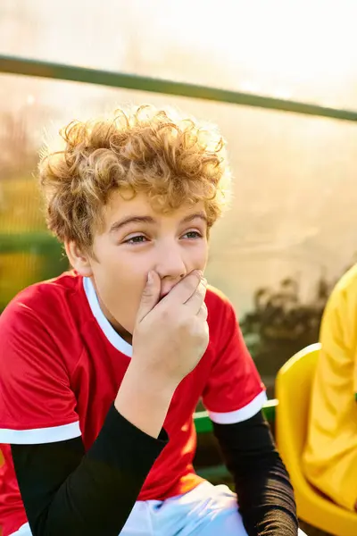 stock image A young boy with a pensive expression sits on a bench, his hand gently placed on his mouth.