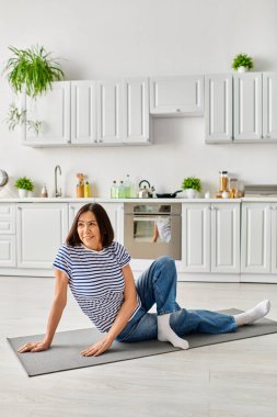 Rahat ev kıyafetleri içinde olgun bir kadın mutfağındaki bir paspasla yoga yapıyor..