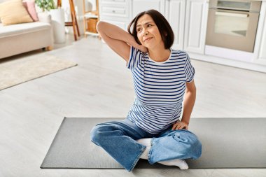 Mature woman in cozy homewear practicing yoga on a mat in her living room. clipart
