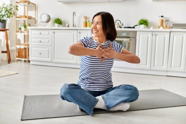 Rahat ev kıyafetleri içinde olgun bir kadın mutfağındaki bir paspasla yoga yapıyor..