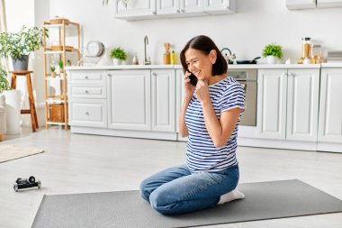 Evde oturan olgun bir kadın yoga minderinde otururken telefonuyla sohbet ediyor..