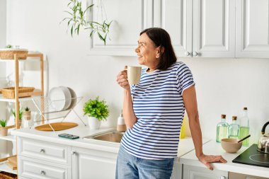 A mature woman enjoys a serene moment in her kitchen, holding a cup of coffee. clipart