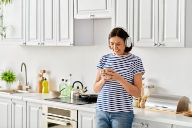 Konforlu ev elbiseli kadın mutfakta duruyor, cep telefonu ekranına dalmış..
