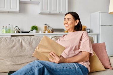 A mature woman in homewear sits on a couch, holding a brown paper bag. clipart