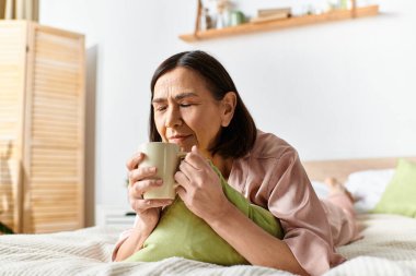 A woman in cozy homewear sitting on a bed, holding a cup of coffee. clipart