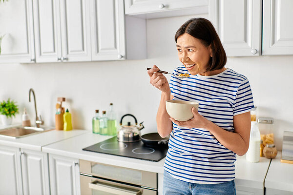 A mature woman in cozy homewear enjoys a bowl of cereal in her kitchen.