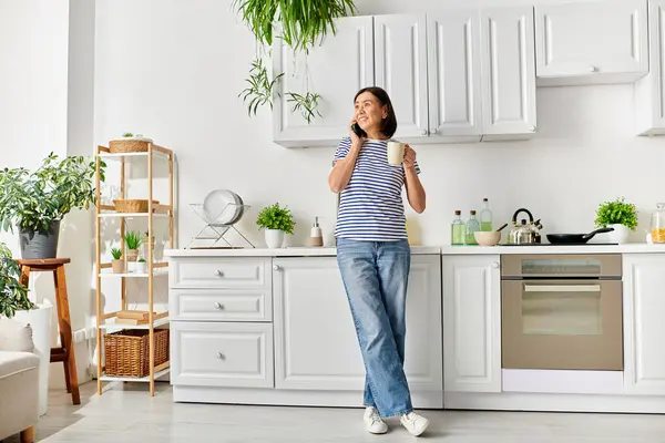 stock image A mature woman in cozy homewear enjoying a moment in her kitchen, holding a cup.