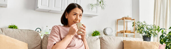 A mature woman in cozy homewear sitting on a couch, gracefully sipping from a cup.