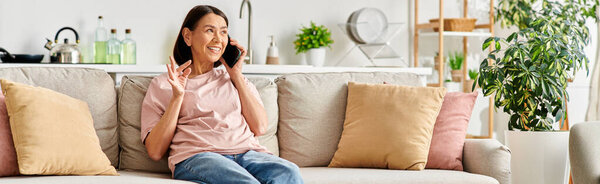 A mature woman in homewear sits on a couch, engaged in a phone conversation.