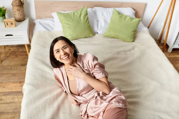 Mature woman relaxes on pink bed in cozy robe.