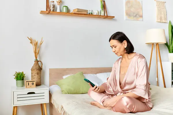 stock image A mature woman in cozy homewear sitting on a bed, engrossed in her phone.