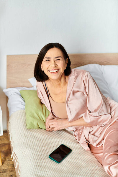 A mature woman in cozy homewear sits on a bed next to a phone.