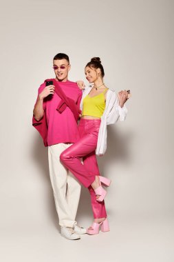 Pembe elbiseli kadın ve şık bir erkek yan yana durur. Çift, gri arka planlı bir stüdyo ortamında aşk ve moda sergiliyor..