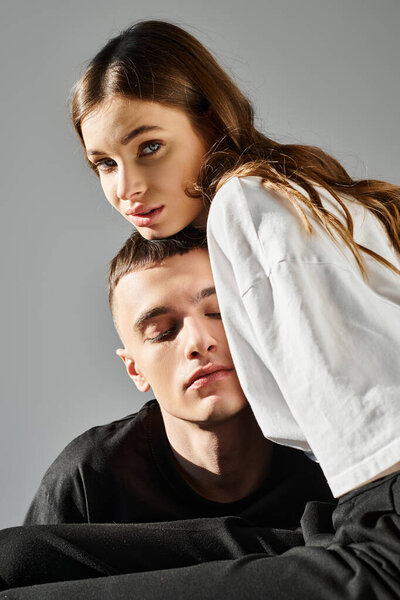 A man sitting beside a woman who is playfully covering her eyes with her hands, both smiling in a studio with a grey background.