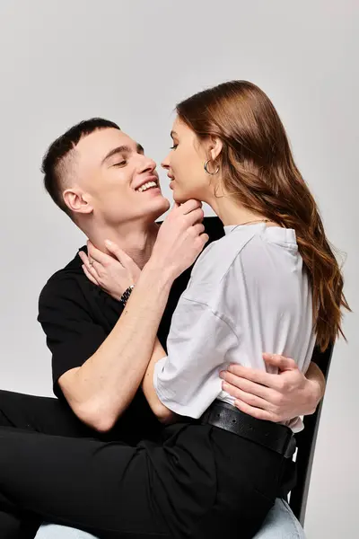 stock image A young man and woman sit closely together on a chair in a studio setting against a grey background.