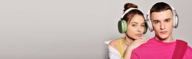 Bir erkek ve bir kadın, ikisi de şık giyinmiş, müzik dinliyorlar. Stüdyoda kulaklıkları takıyorlar. Gri bir arka planı var..