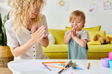 Kıvırcık bir anne, yürümeye yeni başlayan kızının yanında durmuş evde Montessori eğitimini kucaklıyor..