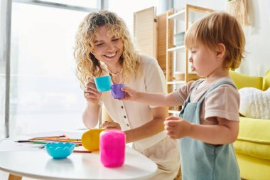 Kıvırcık saçlı bir kadın, yürümekte olan kızıyla masada etkileşime geçiyor ve Montessori metodu aktiviteleriyle meşgul oluyor..
