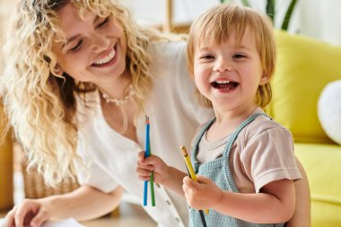 Kıvırcık saçlı bir anne ve küçük kızı ellerinde kalemle evde Montessori eğitim yöntemini uyguluyorlar..
