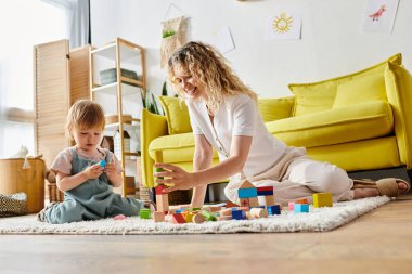 Kıvırcık bir anne, yürümeye yeni başlayan kızını yerde Montessori aktiviteleriyle meşgul ediyor..