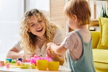Kıvırcık saçlı bir kadın, yürümekte olan kızıyla masada Montessori yöntemlerini kullanır..