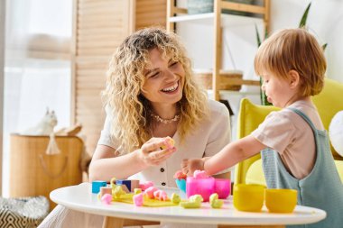 Kıvırcık saçlı bir anne ve yürümeye yeni başlayan kızı Montessori eğitim yöntemini kullanıyorlar, masada birlikte oynuyorlar..