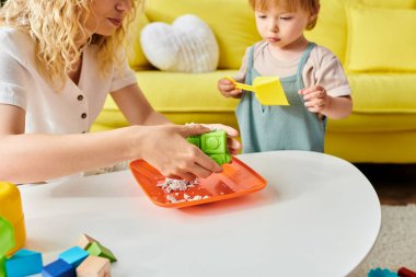 Kıvırcık saçlı bir anne, küçük kızıyla Montessori oyununda karşılaşır, rahat oturma odalarında öğrenme ve eğlenmeyi teşvik eder..