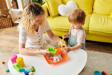 Kıvırcık saçlı bir anne ve küçük kızı evde oyuncaklarla oynayarak Montessori metoduna giriyorlar..