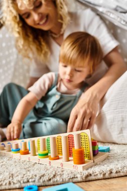Yerde kıvırcık saçlı bir anne, küçük kızıyla Montessori eğitim yöntemlerini kullanarak oynuyor..