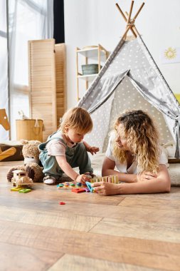 Kıvırcık saçlı bir anne ve küçük kızı yerde oyuncaklarla oynayıp Montessori eğitim metodunu uyguluyorlar..