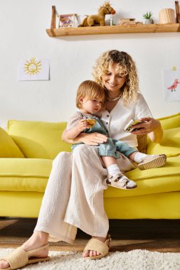 Kıvırcık saçlı bir anne sarı bir kanepede oturur, küçük kızını sıcak ve sevgi dolu bir şekilde kucağına alır..