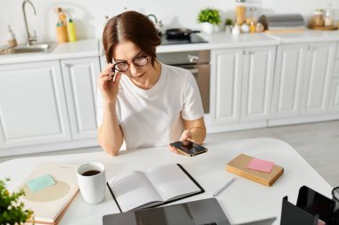 Orta yaşlı bir kadın kendini bir deftere kaptırmış ve bir masada otururken cep telefonunu kontrol ediyor..
