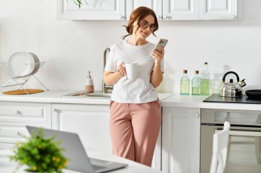 Orta yaşlı bir kadın elinde telefonla mutfakta duruyor..