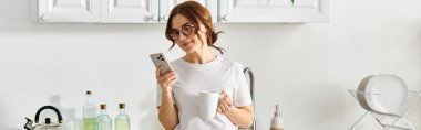 Middle-aged woman standing in kitchen, holding cell phone. clipart