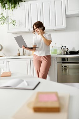 Middle-aged woman stands in kitchen, holding a laptop. clipart