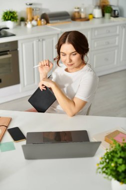 Orta yaşlı bir kadın mutfak masasında dizüstü bilgisayarla çalışıyor..