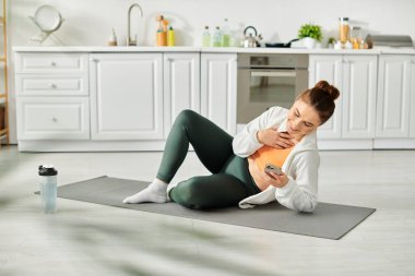 Orta yaşlı bir kadın yoga minderinde oturuyor, elinde cep telefonu var..