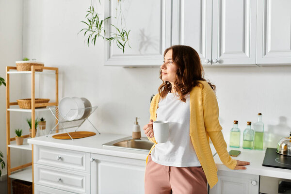 Middle-aged woman calmly standing in kitchen, holding cup.