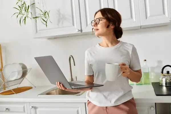 A middle aged woman multitasking with a coffee cup and laptop at home.